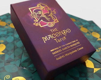 Op de afbeelding: Een paarse doos met gouden letters die "The Macondo Tarot" lezen en is geïnspireerd door de personages van Gabriel Garcia Marquez. De doos is omringd door groene en gouden kaarten.