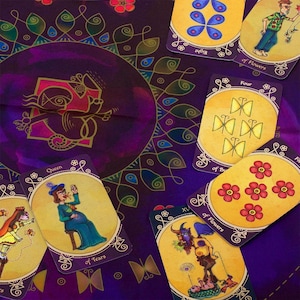 Op de afbeelding: Een tarotkaartendek met een paarse achtergrond en gouden accenten. De kaarten bevatten kleurrijke illustraties van vlinders, bloemen en mensen. De kaarten zijn in een cirkel gerangschikt rond een centrale kaart met een paarse achtergrond en gouden accenten. De centrale kaart bevat een gestileerde afbeelding van een vogel.