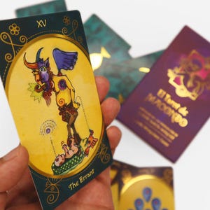 Op de afbeelding: Een tarotkaart met een gouden rand en een gele achtergrond. De kaart toont een geel en rood wezen met vleugels en hoorns dat op een groen en geel platform staat. Het wezen houdt een baby in zijn armen. De kaart is gelabeld met "The Errant" en "XV".