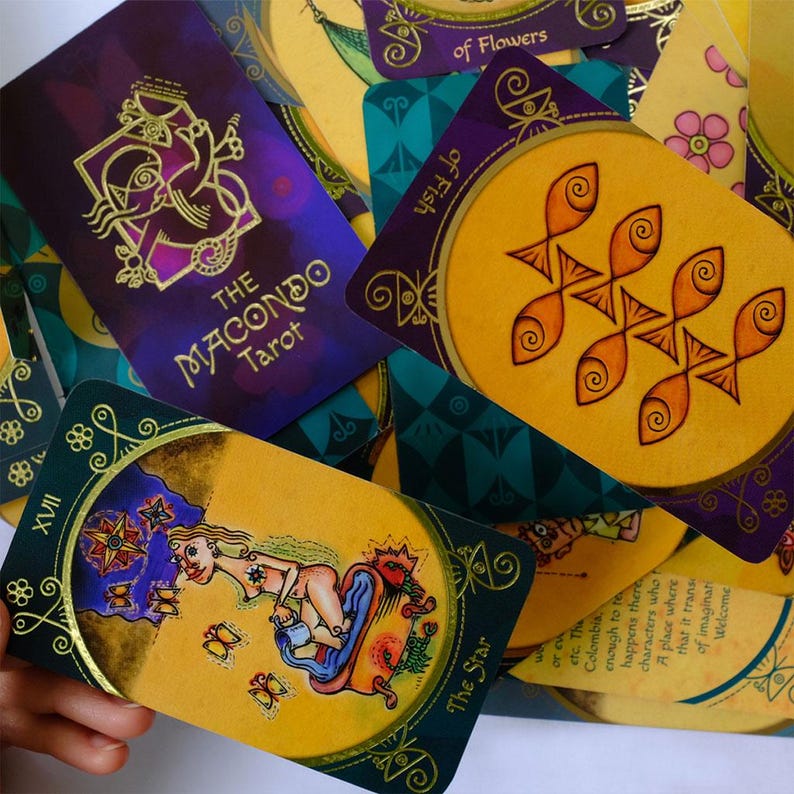 Op de afbeelding: Een set tarotkaarten met kleurrijke illustraties en gouden accenten. De kaarten bevatten verschillende afbeeldingen, waaronder een vrouw in bad, een ster en een vis. De kaarten maken deel uit van het Macondo Tarot-deck.