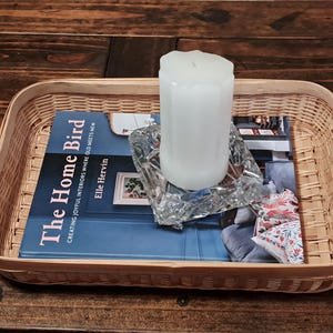 May include: A rectangular woven basket holds a book titled "The Home Bird" and a white pillar candle. The book is on top of a magazine, and the candle sits on a glass ashtray. The basket is on a dark wood surface.