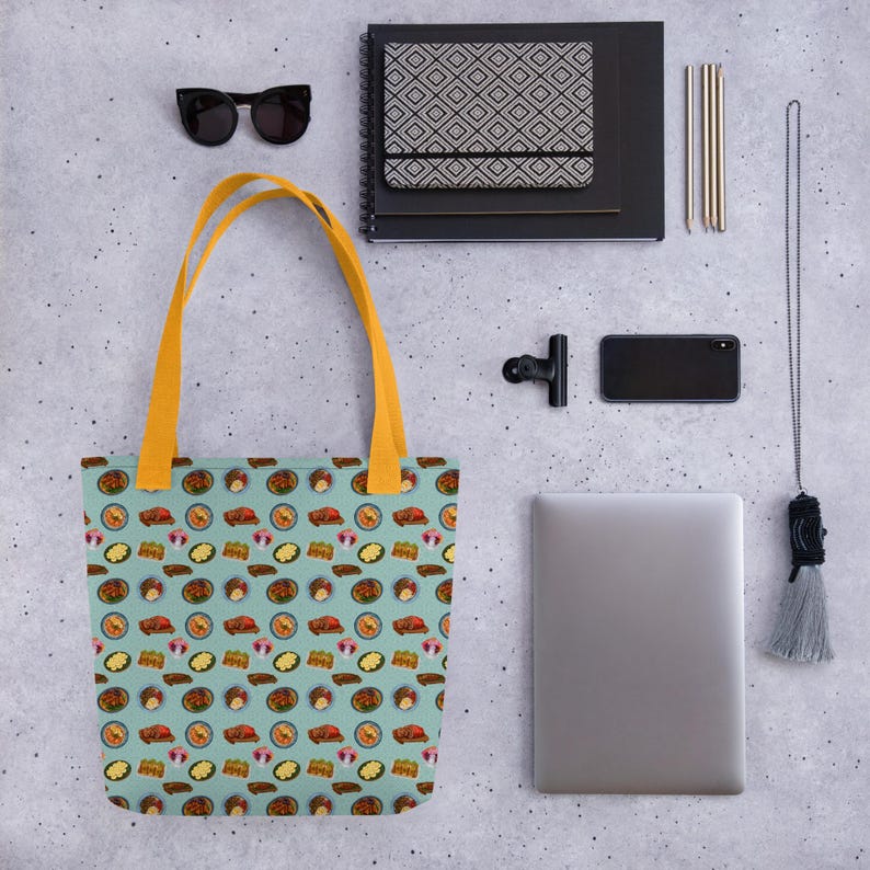 Pu&ograve; includere: Una borsa tote blu con un manico giallo e un motivo di illustrazioni di cibo colorate. La borsa &egrave; su una superficie grigia con altri oggetti, tra cui un laptop, occhiali da sole e un taccuino.