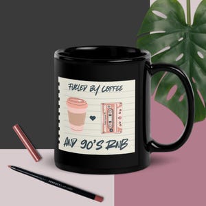 May include: Black coffee mug with a white graphic that says "Fueled by coffee and 90's R&B". The graphic includes a pink coffee cup and a pink cassette tape.