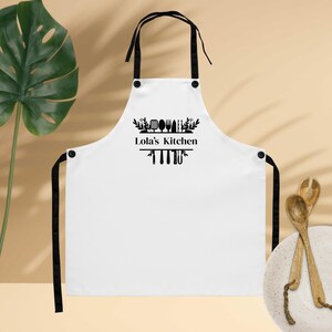 May include: White apron with black straps and trim. The apron features the text "Lola's Kitchen" in black, along with a decorative design of kitchen utensils. The apron is designed for use in the kitchen.