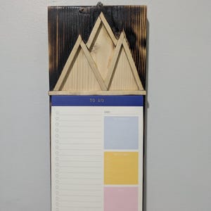 May include: A wooden to-do list board with a burnt wood finish and a mountain design. The board has a white notepad with a blue header that says "TO DO" and a date section. The notepad has three colored sections for writing notes.