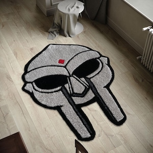 May include: A gray and black rug in the shape of a mask, with a red square accent. The rug has a textured surface and is placed on a wooden floor. The mask design features large black eye shapes and a detailed outline.