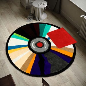Pode incluir: Um tapete redondo com um design colorido que se assemelha a um disco de vinil. O tapete apresenta uma borda preta, um centro cinza e vermelho, e segmentos radiais em várias cores, incluindo branco, azul, verde e laranja. Um pequeno tapete retangular vermelho está em cima.
