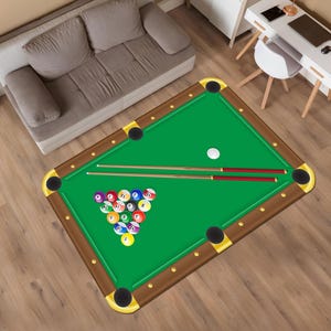 May include: A rectangular green pool table rug with brown trim and gold corner pockets. The rug features a rack of numbered billiard balls, two pool cues, and a white cue ball. The rug is on a wooden floor.