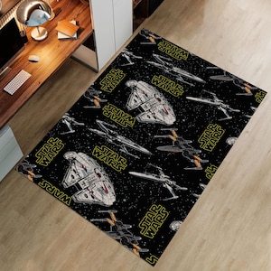 Puede incluir: Alfombra rectangular negra con un diseño de Star Wars. La alfombra muestra ilustraciones de naves espaciales, como el Halcón Milenario y cazas X-wing, sobre un fondo estrellado. Las palabras "STAR WARS" están impresas en amarillo.
