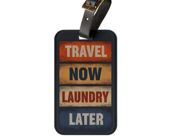 Op de afbeelding: Een rechthoekig bagagelabel met een zwarte rand en een zwarte leren riem met een goudkleurige metalen gesp. Het label toont vier horizontale panelen met de tekst TRAVEL in oranje, NOW in donkerblauw, LAUNDRY in rood en LATER in lichtblauw.