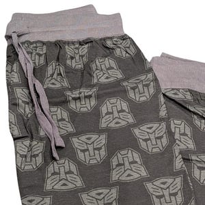 May include: Dark gray pajama pants with a repeating gray Transformers logo pattern. The waistband and drawstring are gray. The pants are made of a soft, comfortable material.
