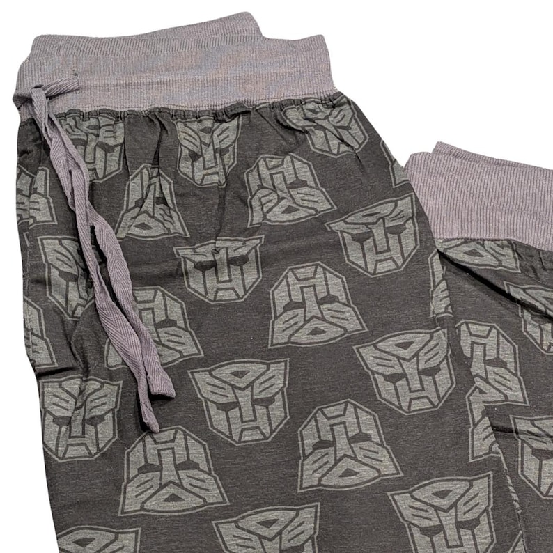 May include: Dark gray pajama pants with a repeating gray Transformers logo pattern. The waistband and drawstring are gray. The pants are made of a soft, comfortable material.