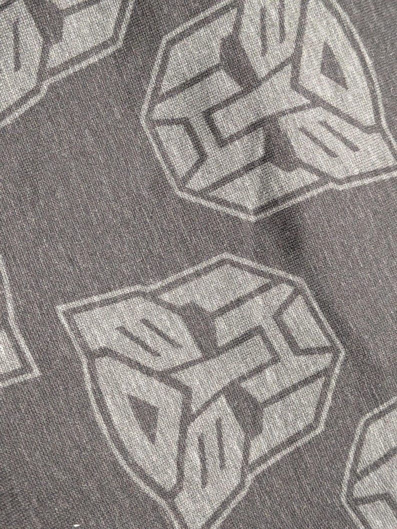 May include: Close-up of a gray fabric with a repeating pattern of a stylized, geometric emblem in a lighter gray. The emblem has a hexagonal shape with angular lines and shapes, creating a textured appearance.