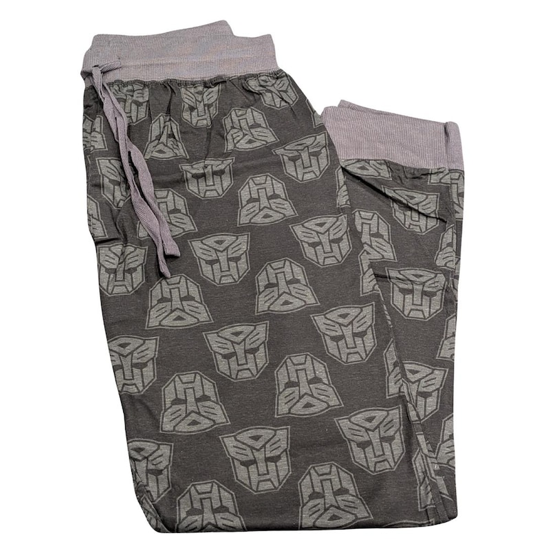 May include: Gray pajama pants featuring a repeating pattern of gray Transformers Autobot symbols. The pants have a gray drawstring waistband and gray cuffs. The fabric appears to be soft and comfortable, suitable for lounging or sleeping.