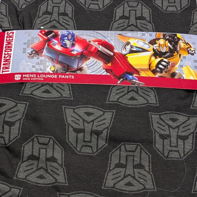 May include: Dark gray lounge pants with a repeating pattern of Transformers logos. The packaging features images of Optimus Prime and Bumblebee. The text on the packaging reads "Transformers" and "Mens Lounge Pants 100% Cotton."