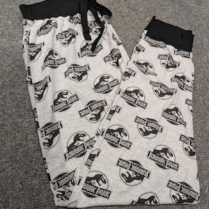 May include: Gray pajama pants with a black waistband and drawstring. The pants are patterned with the "Jurassic Park" logo and a dinosaur silhouette in black. The pants are folded on a gray surface.
