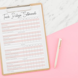 May include: A clipboard with a weekly cleaning schedule in Italian, featuring pink and white boxes for tracking tasks. A white pen with gold accents rests on a pink surface, next to the clipboard. The background is a mix of pink and white.