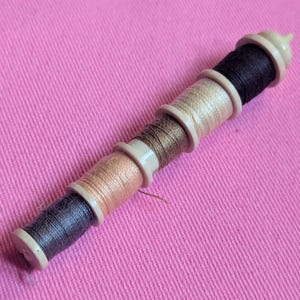 May include: A multi-spool thread holder with various colors of thread, including black, tan, and brown. The spools are held together by a beige plastic casing. The background is a solid pink fabric.
