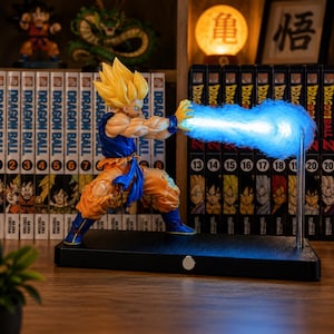 Peut inclure: Une figurine articulée de Super Saiyan Goku dans une pose de combat dynamique, portant un gi bleu et orange, avec un effet d'explosion d'énergie LED bleu brillant s'étendant des mains, montée sur une base rectangulaire noire avec une tige.