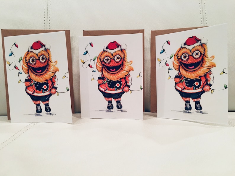 May include: Three Christmas cards featuring a cartoon mascot wearing a Santa hat and holding a string of Christmas lights. The mascot is wearing an orange and black hockey jersey with a logo on the front.