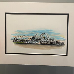 May include: Watercolor painting of a pier with a Ferris wheel, roller coaster, and other amusement park rides. The artwork is framed with a white mat and black frame. The sky is a mix of blue, yellow, and orange.