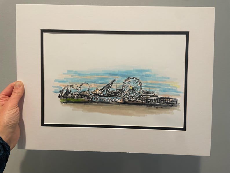 May include: Watercolor painting of a pier with a Ferris wheel, roller coaster, and other amusement park rides. The artwork is framed with a white mat and black frame. The sky is a mix of blue, yellow, and orange.
