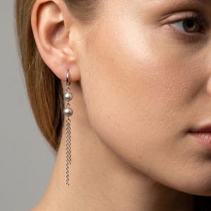 May include: A silver-toned hoop earring featuring two smooth, round, grey beads suspended vertically. Below the beads, three delicate, thin silver-toned chains hang down, creating a layered, dangling effect that catches the light.