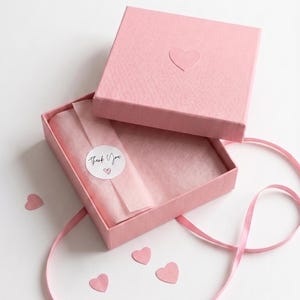 Op de afbeelding: Een vierkante roze geschenkdoos met een textuurafwerking en een klein reli&euml;fhart op het deksel. De open doos bevat lichtroze vloeipapier dat is vastgezet met een ronde sticker met de tekst Thank You en een hartje. Roze lint en papieren hartjes.