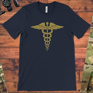 May include: Navy blue t-shirt featuring a gold-colored caduceus symbol, a staff with two snakes and wings. The shirt is made of a soft material. The shirt is displayed on a wooden surface, with military-style clothing and accessories in the background.