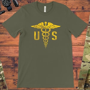 Może przedstawiać: Zielony T-shirt z żółtym amerykańskim symbolem medycznym i literami "US" nadrukowanymi na przodzie.