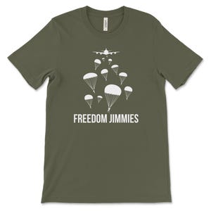 May include: Olive green t-shirt with a white graphic of an airplane and parachutes. The text "FREEDOM JIMMIES" is printed in white below the graphic. The shirt is made of a soft material.