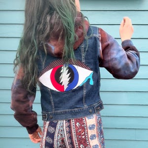 May include: A dark wash denim jacket with a tie-dye hood and sleeves. The back of the jacket features a large, colorful eye patch with a lightning bolt design. The jacket has a button-up front and pockets.