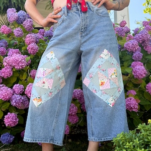 May include: Light blue denim jeans featuring a wide-leg cropped silhouette and a high-waisted fit. Each leg displays a large, diamond-shaped patchwork panel constructed from various floral and geometric patterned fabric squares in pastel colours.