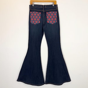 Puede incluir: Vaqueros acampanados de mezclilla azul oscuro con bolsillos con estampado de rosquillas rojas. Los vaqueros cuelgan de una percha de madera sobre un fondo blanco. Los bolsillos son de un color contrastante y a&ntilde;aden un elemento de dise&ntilde;o &uacute;nico.