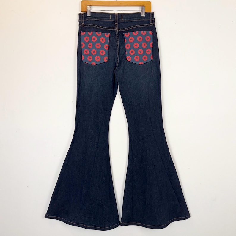 Puede incluir: Vaqueros acampanados de mezclilla azul oscuro con bolsillos con estampado de rosquillas rojas. Los vaqueros cuelgan de una percha de madera sobre un fondo blanco. Los bolsillos son de un color contrastante y a&ntilde;aden un elemento de dise&ntilde;o &uacute;nico.