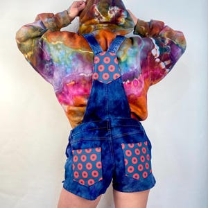May include: A pair of blue denim overalls with red and blue donut print on the back pockets. The overalls are worn over a tie-dye hoodie.