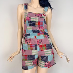 Può includere: Salopette corta patchwork multicolore. I pantaloncini presentano un mix di quadrati rosa, blu, rossi e blu navy. Sono visibili spalline regolabili con hardware in metallo. Il capo è realizzato con un patchwork di tessuti fantasia.