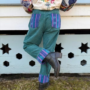 May include: Teal corduroy pants with patterned purple and blue pocket and cuff accents. The pants are paired with black leather boots. The person is wearing a cream-colored jacket with floral embroidery and patchwork sleeves.