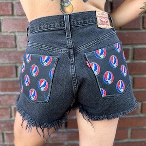 Könnte beinhalten: Schwarze Jeansshorts mit ausgefransten Kanten. Die Gesäßtaschen sind mit einem sich wiederholenden Muster des Steal Your Face-Logos der Grateful Dead in Rot, Blau und Weiß auf schwarzem Grund verziert. Ein Levi Strauss & Co. Etikett ist sichtbar.