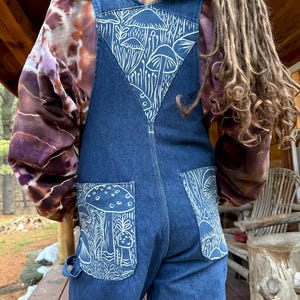 May include: Dark blue denim overalls with white mushroom and plant designs on the back and pockets. The overalls have a bib front and adjustable straps. The person is wearing a tie-dye hoodie underneath.