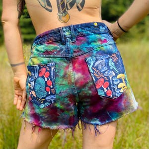 May include: Denim shorts with a vibrant tie-dye pattern in shades of blue, green, and pink. The back pockets feature a dark blue fabric with a colorful mushroom and plant design. The shorts have a frayed hem.