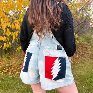 Op de afbeelding: Lichtblauwe denim tuinbroekshorts met een witte madeliefjesprint. De achterzakken zijn voorzien van een rood en donkerblauw bliksemschichtmotief. Een kleine zwarte rechthoekige patch met witte tekst is zichtbaar op het bovenste rugpand.