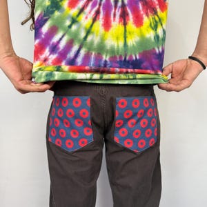 May include: Dark brown pants with two back pockets featuring a blue fabric with a red donut pattern. The person is wearing a tie-dye shirt with a mix of colors including purple, green, yellow, and red.