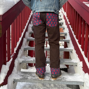 Könnte beinhalten: Braune Hose mit Taschen und Manschetten mit blauem Hintergrund und rotem Blumenmuster. Die Person trägt eine Jeansjacke und schwarze Stiefel. Die Person steht auf einer schneebedeckten Metalltreppe mit roten Geländern.