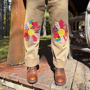 May include: Tan trousers featuring large, circular knee patches made from a patchwork of colorful fabric scraps in various patterns, including floral, polka dot, and geometric designs, with brown leather lace-up boots worn on the feet.