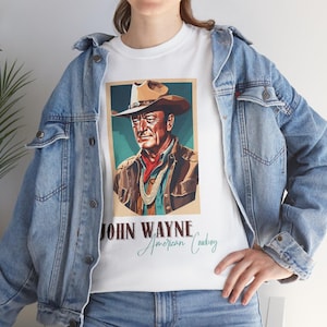 Puede incluir: Camiseta blanca con un retrato de John Wayne con sombrero de vaquero y el texto "JOHN WAYNE American Cowboy". La imagen tiene una estética vintage, con una paleta de colores marrón, verde y rojo.