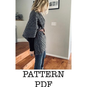 Op de afbeelding: Een grijze gebreide cardigan met een kabelbreipatroon en een bruine leren riem. De cardigan heeft een wijde, vloeiende silhouet en wordt gedragen door een persoon die op een houten trap staat. De tekst "PATTERN PDF" is onder de afbeelding gedrukt.