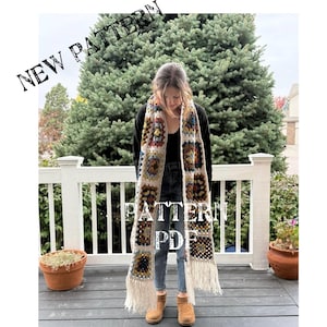 May include: A white crochet scarf with colorful granny squares and fringe. The scarf is worn by a person standing on a wooden deck.