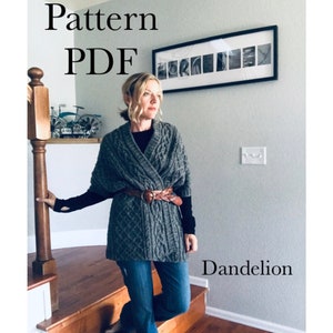 Pode incluir: Um envoltório de malha cinzento com um padrão de malha de cabo e um cinto de couro castanho. O texto "Pattern PDF" e "Dandelion" é visível na imagem.