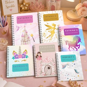 May include: A collection of spiral-bound notebooks with whimsical designs, including a unicorn, fairy, and castle. Each notebook features the text "Katrina's Drawings and Doodles" on the cover. The notebooks are arranged on a pink surface with decorative elements.